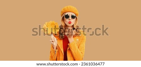 Autumn color style outfit, portrait stylish beautiful young woman model with yellow maple leaves blowing her lips sends sweet kiss wearing orange french beret, sunglasses on beige studio background