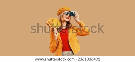 Autumn color style outfit, beautiful young woman photographer with film camera holds yellow maple leaves wearing orange french beret hat, jacket on beige studio background
