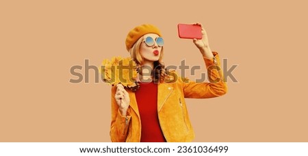 Autumn style outfit, stylish modern young woman taking selfie with mobile phone holds yellow maple leaves wearing orange french beret hat, jacket and round sunglasses on beige studio background