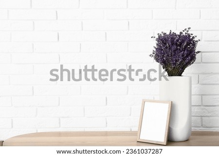 Vase with beautiful lavender flowers and blank frame on table near light brick wall in room, closeup