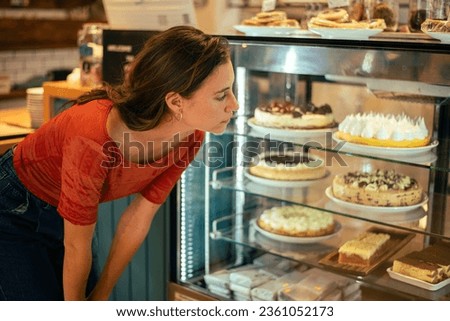 Beautiful 25-year-old woman, white with freckles, wearing jeans and a red t-shirt, in a specialty café, placing her order, choosing a sweet cake to eat. She's in a modern café Royalty-Free Stock Photo #2361052173