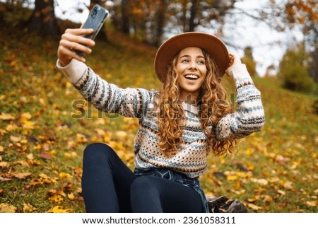 Beautiful woman in a stylish sweater with a phone in her hands sits in a clearing among yellow fallen leaves in an autumn park. Tourist enjoys the weather, takes a photo with a yellow leaf. 