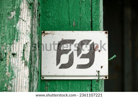 An old, scratched, rusty, paint streaked address sign with the number 69 attached to a nail on the corner of a wooden house with cracked green paint. Selective focus.