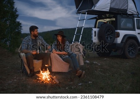 Сouple in love, man and woman on camp in mountains, sit by fire against backdrop of car in mountains.
