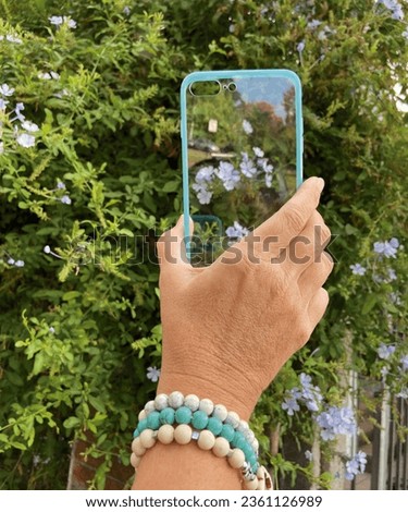 Woman's hand holding a transparent light blue transparent case, over a tree with violet flowers. Where the arm has light blue, beige and white bracelets.