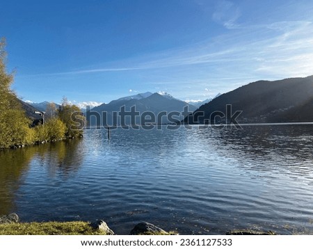 This is a photo-realistic image of a lake with mountains in the background. The lake is calm, the water is a deep blue The sky is blue with wispy clouds The mountains in the background are snow-capped
