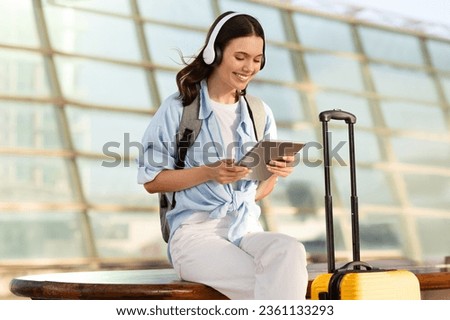 Positive millennial caucasian lady with suitcase in headphones, chatting on tablet at airport, train station. Travel blog, online booking, trip lifestyle, human emotions and music Royalty-Free Stock Photo #2361133293