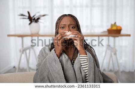 Sick unhappy millennial long-haired black lady sitting on couch covered in warm blanket at home, sneezing, using napkin, suffering from cold, flu or coronavirus, copy space, closeup Royalty-Free Stock Photo #2361133311