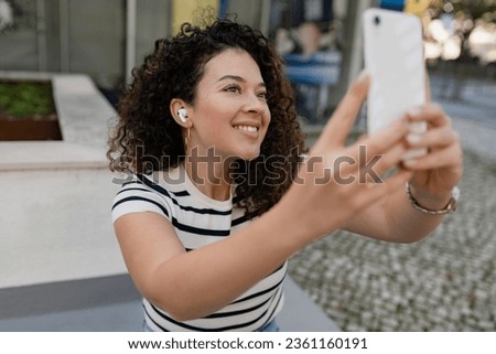 pretty curly woman walking in city street in striped t-shirt, using smart phone, taking photo, making selfie picture