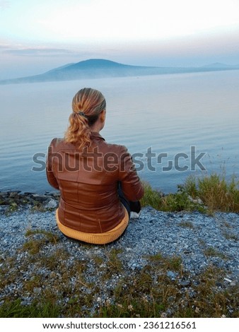 Landscape with grass and blue sky. A girl sits on the shore of a forest lake and looks into the distance