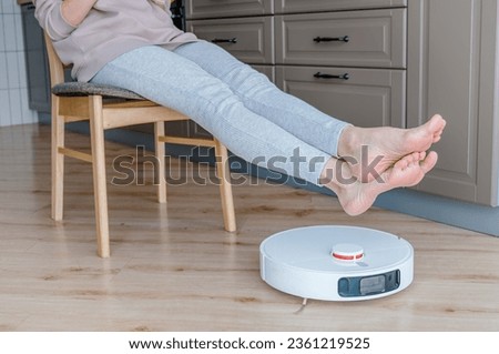 Robot vacuum cleaner cleaning the living room. Young woman enjoy rest, sitting on chair at home
