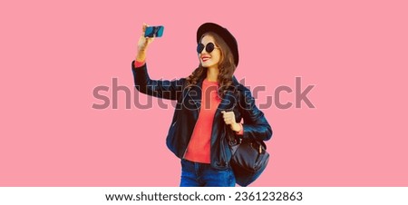 Stylish modern smiling young woman taking selfie with mobile phone wearing black round hat, jacket and backpack on pink studio background