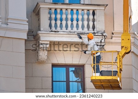 Worker on crane repairing balcony, plastering cracked stucco with trowel, renovation work, restore facade of historic building. Plaster cladding and crack repair. Plasterer repair balcony of building Royalty-Free Stock Photo #2361240223