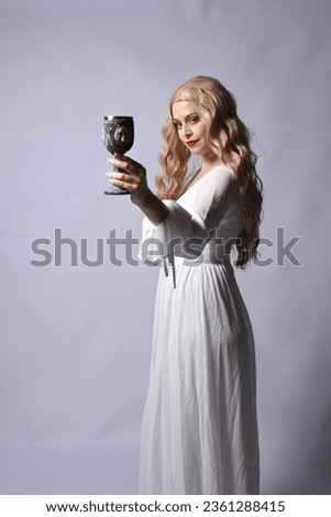 Close up portrait of beautiful blonde model wearing elegant  white halloween gown, a historical fantasy character.  Holding wine goblet, isolated on audio background. Royalty-Free Stock Photo #2361288415