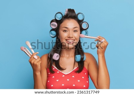 Wonderful Asian girl in red top puts her hair in curlers and uses cosmetics brushes to blend blush, cosmetic procedure concept, copy space
