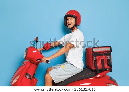 Young Asian courier guy on red scooter with big delivery bag holds hands on handlebar and smiles, picture setup on blue backdrop, fast delivery concept, copy space