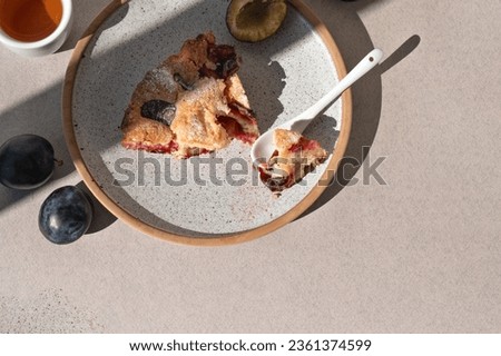 Piece of bitten plum pie on plate with dessertspoon, on beige table background. Delicious fruit bakery, cake with plum, cinnamon and sugar powder.