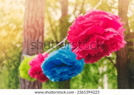 Close up of a colorful party garland made of paper flowers tied between trees in a park at an open air celebration event.