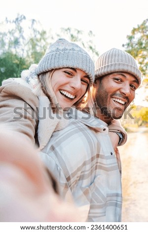 Vertical portrait of a young adult couple having fun together taking a selfie. Carefree boyfriend giving a piggyback ride to his cheerful girlfriend on a weekend sightseeing outside. High quality
