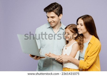 Side view young IT parents mom dad with child kid daughter girl 6 years old wear blue yellow casual clothes hold use work on laptop pc computer isolated on plain purple background. Family day concept