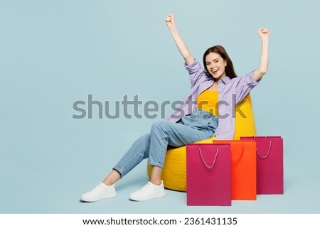 Full body young woman wears casual clothes sit in bag chair near paper package bags after shopping do winner gesture isolated on plain pastel blue cyan background. Black Friday sale buy day concept Royalty-Free Stock Photo #2361431135