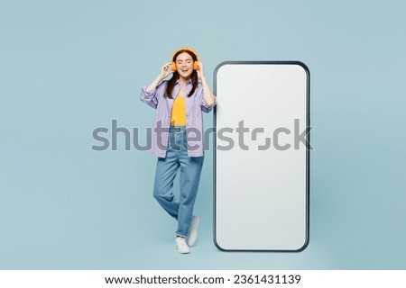 Full body young woman wear purple shirt yellow t-shirt casual clothes big huge blank screen mobile cell phone smartphone with area listen to music in headphones isolated on plain light blue background