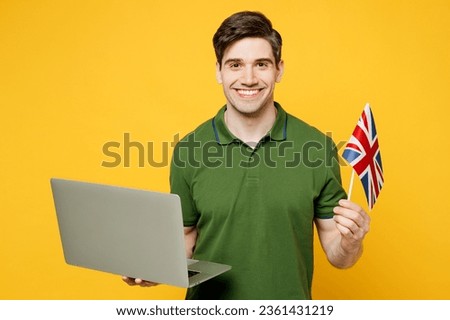 Young caucasian cool happy cheerful IT man wears green t-shirt casual clothes hold British flag use work on laptop pc computer isolated on plain yellow background studio portrait. Lifestyle concept