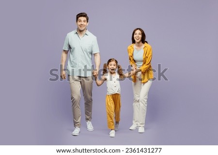 Full body young happy parents mom dad with child kid daughter girl 6 years old wear blue yellow casual clothes hold hands walk going look camera isolated on plain purple background. Family day concept