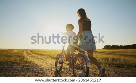 mom teaches son to ride bike. happy family childhood dream concept. mom and little son learn to ride a bike silhouette in the park in sunlight nature. happy family goes in for sports outdoors
