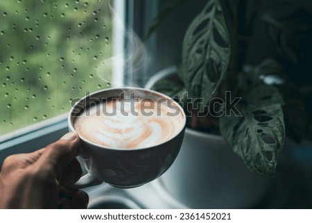 person's hand is holding an aromatic latte art coffee cup. Drink coffee on a rainy day by the window