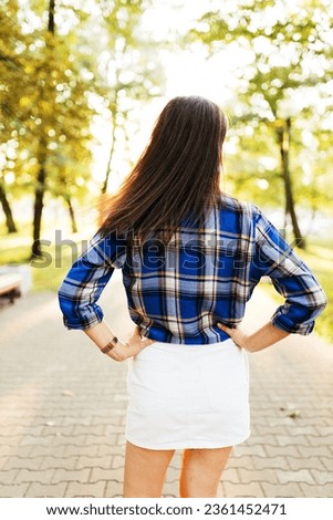 A young beautiful woman in a blue checkered shirt and a white short skirt with black long hair walks and poses in an autumn park. Summer portrait of a beautiful girl on a sunny day