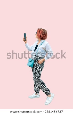 Full length portrait of a pink-haired girl in a white shirt and leopard print trousers taking a selfie with a mobile phone isolated on a pink background
