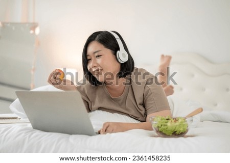 A happy Asian plus-size woman lying on bed, wearing headphones, and using her laptop, watching a movie on bed, reading an online blog, talking on video call, or working from home. Home lifestyle