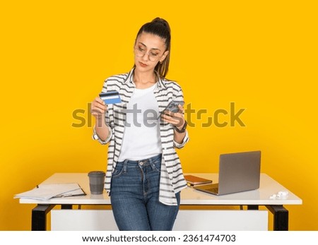 Shopping online via mobile phone application. Business woman leaning office desk holding bank credit card and smartphone, purchase, order, payment concept isolated  yellow studio background. 