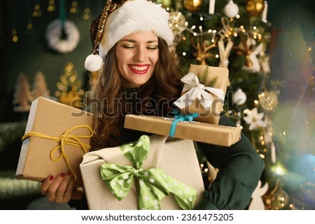Christmas time. smiling elegant housewife with Santa hat and eco packaged present boxes in green dress near Christmas tree at modern home. Royalty-Free Stock Photo #2361475205