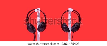 Two pairs of black headphones on stand. Red background, white table and black noise reduction device create creative atmosphere. Individual lecture, listening music, audio training, concern for others Royalty-Free Stock Photo #2361475403