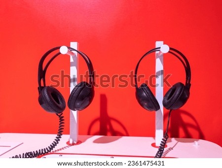 Two pairs of black headphones on stand. Red background, white table and black noise reduction device create creative atmosphere. Individual lecture, listening music, audio training, concern for others Royalty-Free Stock Photo #2361475431