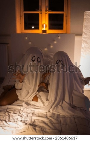 Vertical Foto of two little children play ghost sitting on the bed in the evening in their children's room. Kids dressed in homemade ghost costumes from sheets. Preparing and celebrating the Halloween Royalty-Free Stock Photo #2361486617