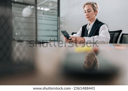 Remote portrait of middle-aged businesswoman using smartphone sitting at office desk, touching screen, typing message on mobile phone, browsing Internet, social media, shopping online.