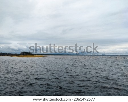 The dark gray waters of the Gulf of Finland merge with light gray clouds.  On the left side of the photo is an island of land with reeds.  Nuclear power plant cooling towers smoke on the horizon.