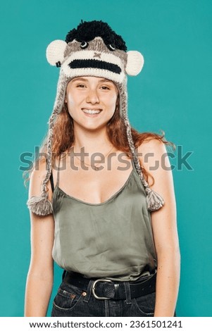 Happy smiling young girl with ginger curly hair and wool beanie standing in studio on blue background and looking at camera.