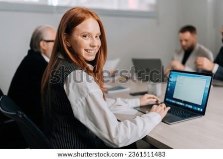 Portrait of attractive secretary smiling looking to camera during meeting with colleagues working on background. Positive redhead businesswoman working typing on laptop computer on boardroom.