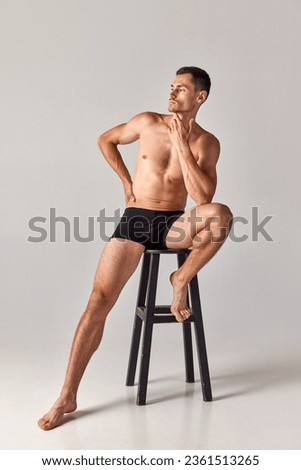 Full-length portrait of handsome, muscular young man sitting on chair in underwear and posing against grey studio background. Concept of men's health and beauty, body care, fitness, wellness, ad Royalty-Free Stock Photo #2361513265
