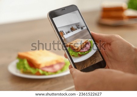 Young woman with mobile phone taking picture of tasty sandwich in kitchen, closeup