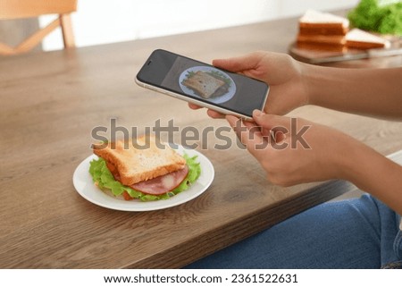 Young woman with mobile phone taking picture of tasty sandwich in kitchen, closeup