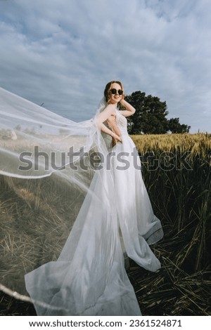 A beautiful young blonde bride in a white dress with a long veil in sunglasses stands in a wheat field outdoors in Ukraine. Wedding photography, portrait.
