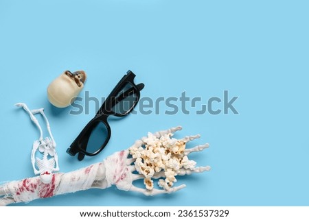 Composition with tasty popcorn, 3D glasses, skeleton and skull for Halloween celebration on blue background
