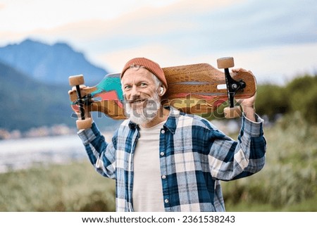 Active cool happy bearded old hipster man standing in nature park holding skateboard. Mature traveler skater enjoying freedom spirit and extreme sports hobby on mountains background. Portrait