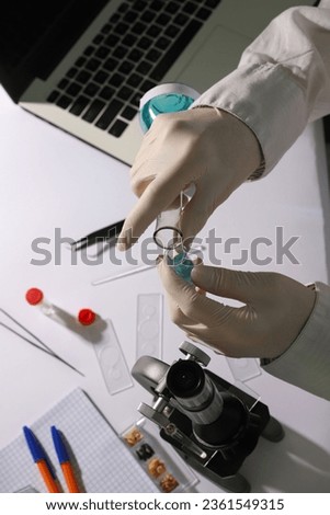 laboratory. on a white table there is a white laptop, test tubes, a microscope. hands in medical gloves hold two test tubes with blue liquid.