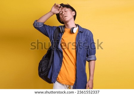 Exhausted Asian man with headphones on neck and carrying backpack touching his head showing tired expression